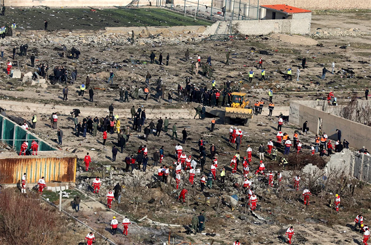Débris d’avion dispersés dans un champ. Un grand nombre de personnes se déplacent parmi les décombres. Un terrain de soccer partiellement détruit occupe la partie supérieure, et un bulldozer est à l’œuvre.