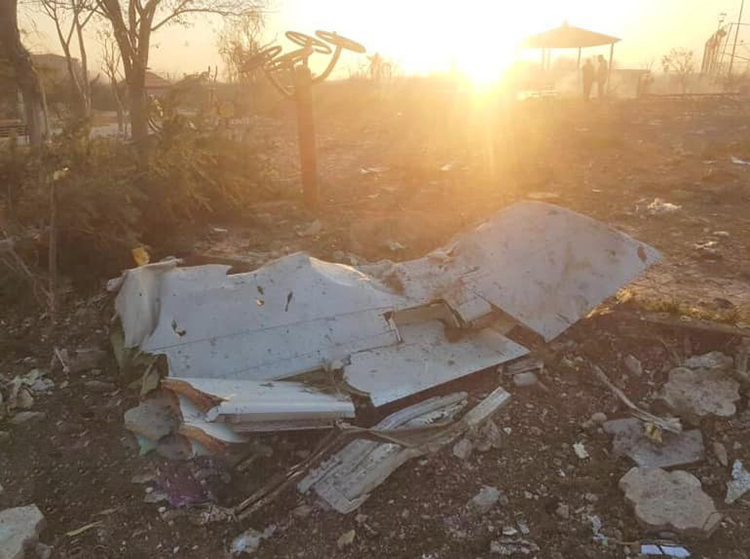 Crumpled portion of the wing from PS752 surrounded by debris on a field. The sun sets in the background.