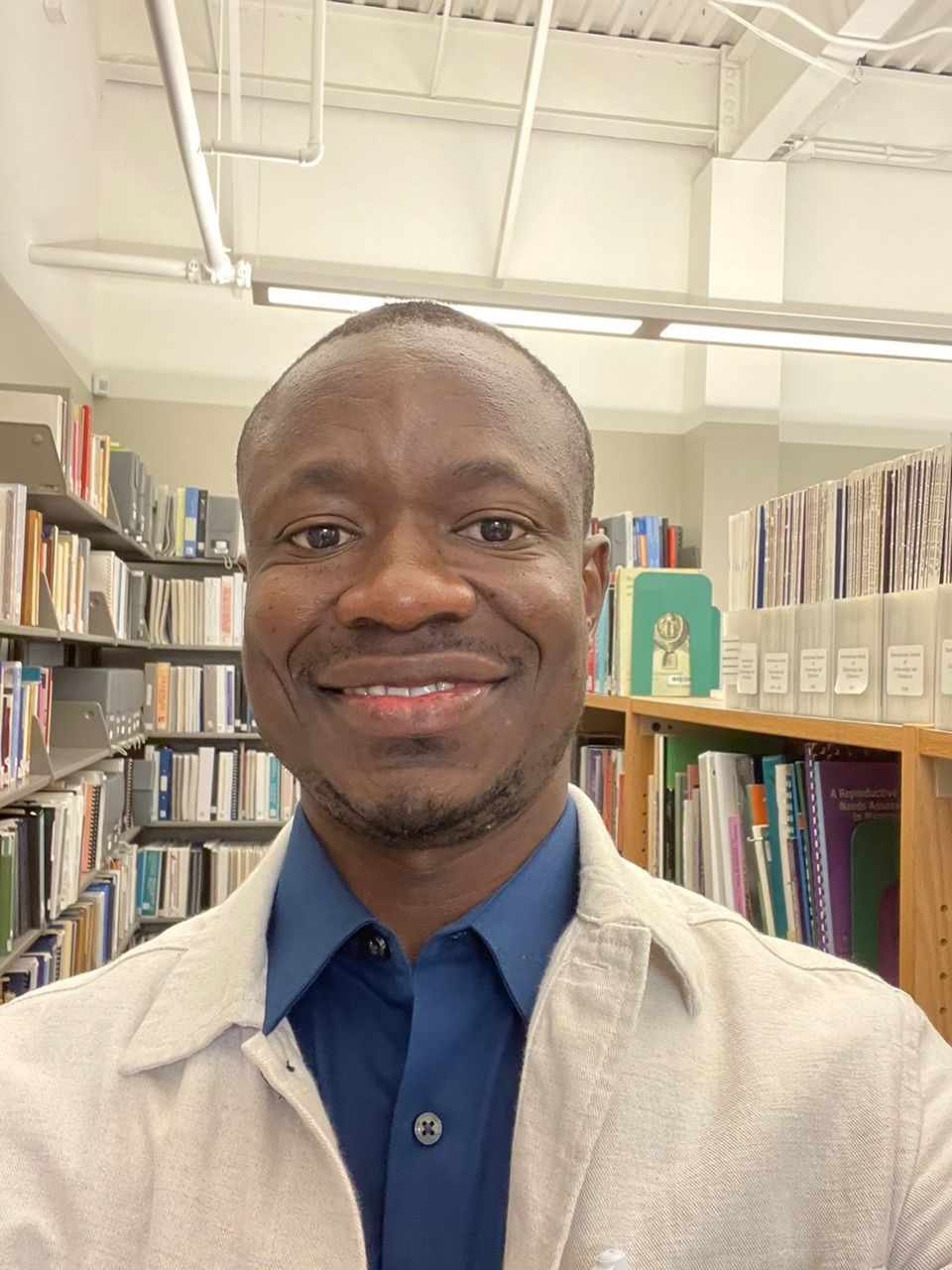 Jean-Claude Mulunda, portant une chemise à col bleu et un veston blanc, sourit à l’intérieur d’une bibliothèque.