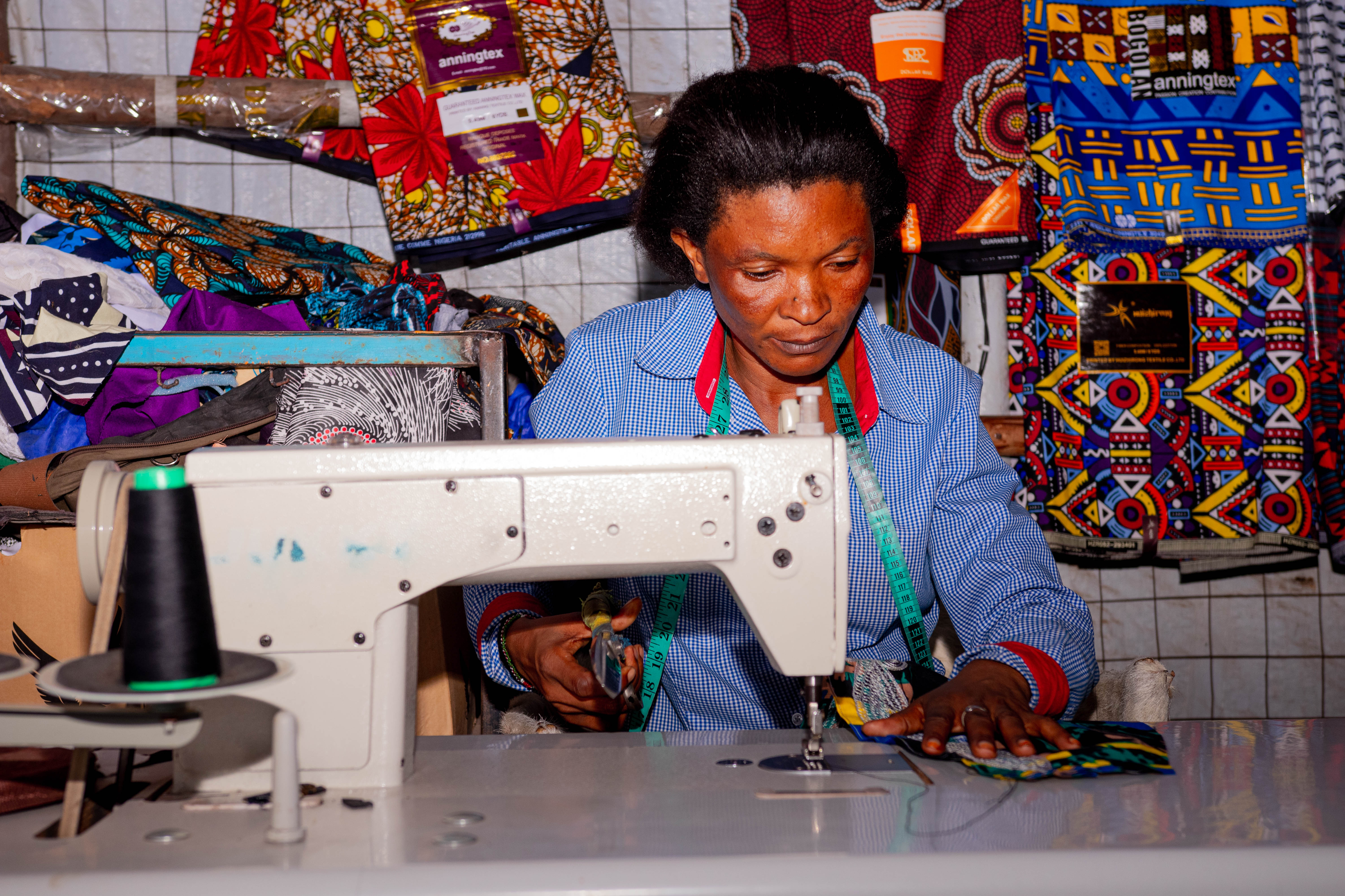 : Adelle uses a sewing machine at her workstation. In the background are various textiles in colourful patterns and designs.