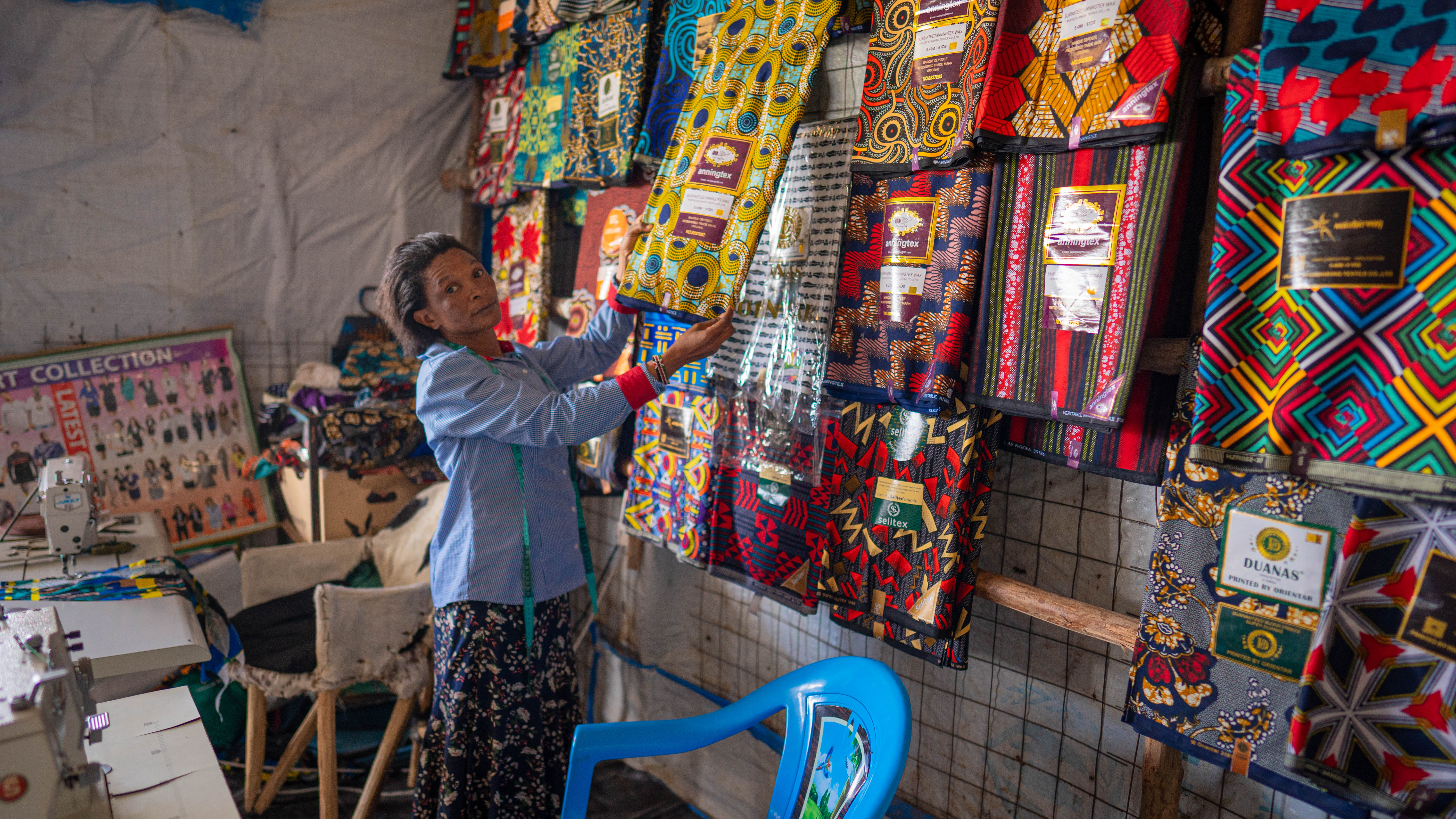Adelle holds up a yellow and blue piece of fabric. She is standing in front of a wall covered in various textiles with colourful patterns and designs.
