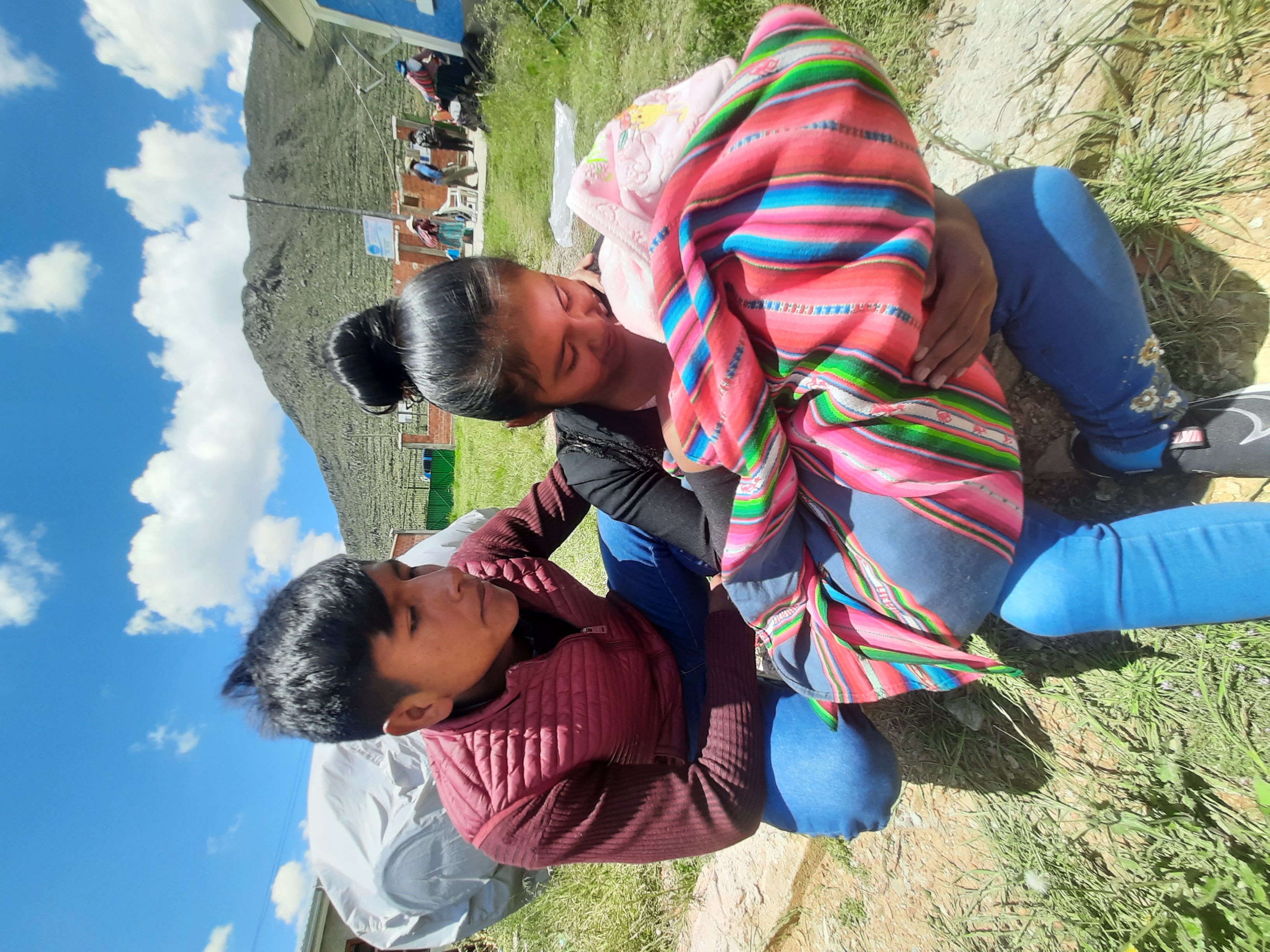 An adolescent girl holds a bundled baby in her arms outdoors. An adolescent boy crouches next to her.