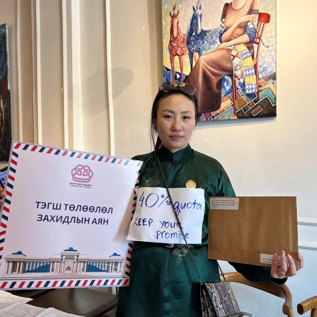 A Mongolian woman wearing a green shirt holds up signs and envelopes during a press conference by the Women&rsquo;s Leadership Network in Mongolia. One sign in Mongolian reads &ldquo;Equal Representation Letter Campaign&rdquo; while another sign strapped to her chest reads &ldquo;40% quota Keep your promise.&rdquo;