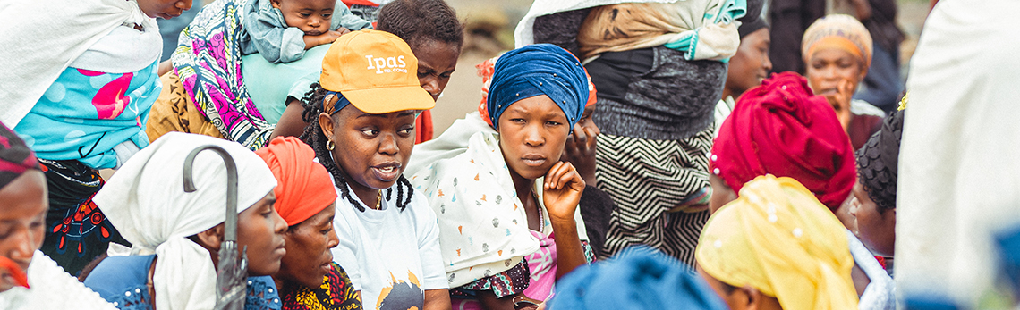 A large group of women and children gather closely together outdoors. Many of the women are wearing colourful headscarves and traditional clothing and some of them are carrying children. Photo credit: Ipas DRC